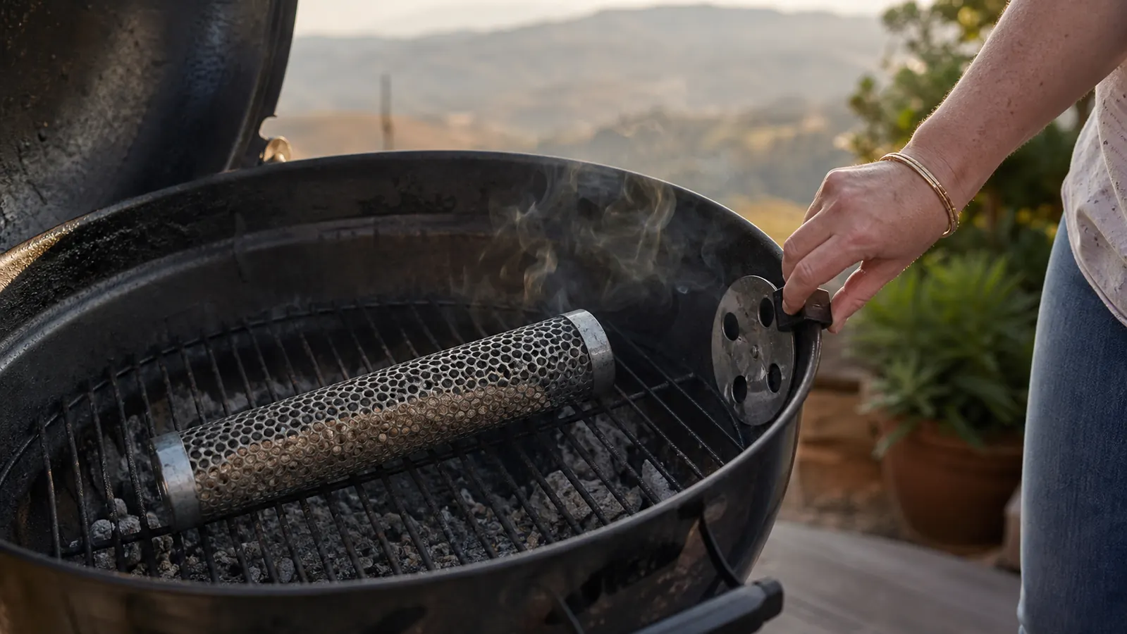 A mesh smoker tube filled with wood pellets resting inside a charcoal kettle grill on a backyard deck at golden hour